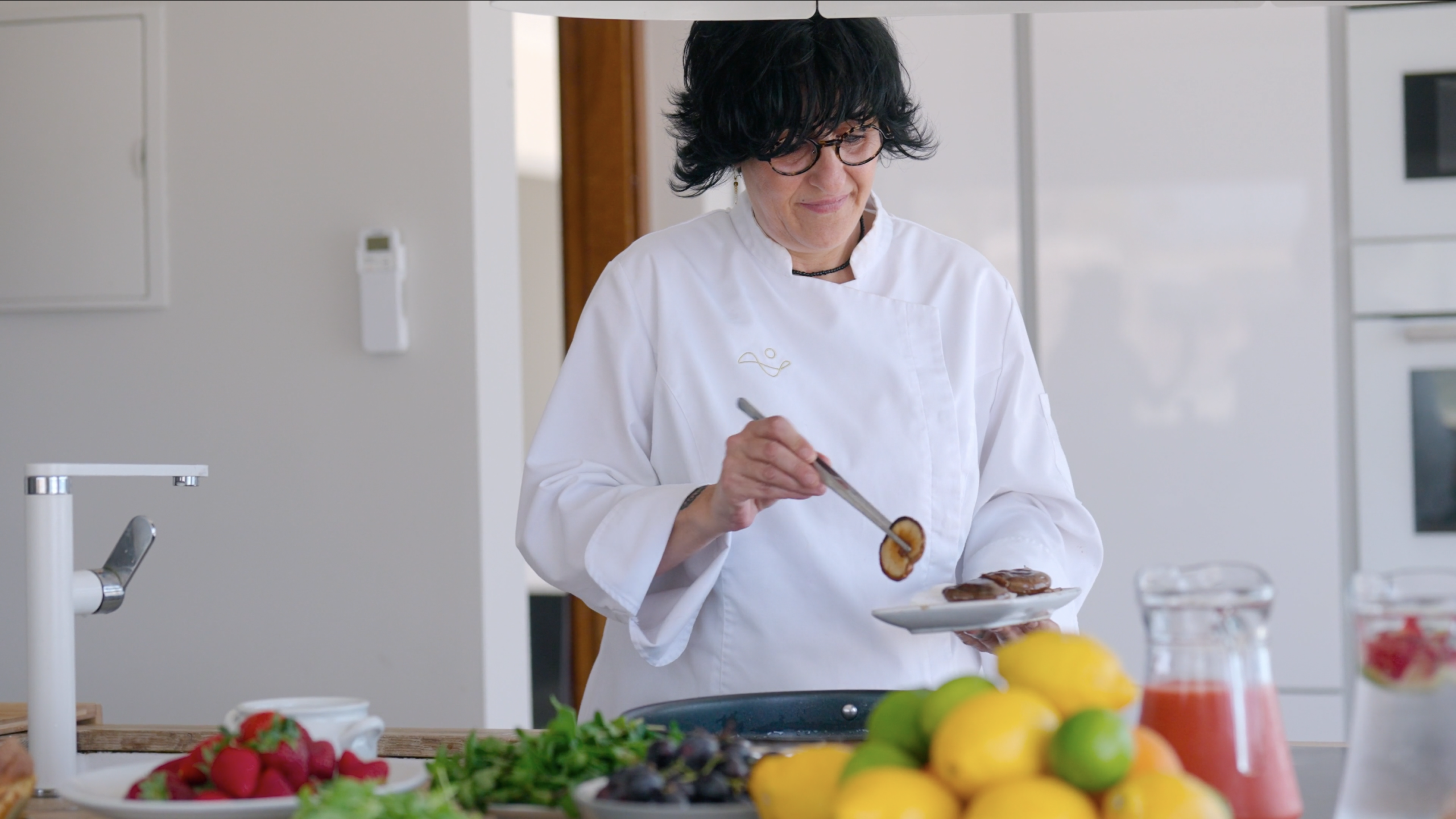 Chef plating food in a bright kitchen with fresh fruit and herbs